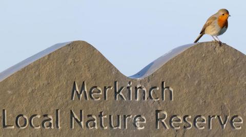 A robin is perched on a stone sign for the reserve. Carved into the stone are the words: "Merkinch Local Nature Reserve". The sky behind the bird and sign is blue. The robin is a small brown bird with a red breast.