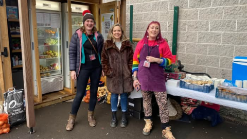 Three women including Vix Ross, right, stand in front of two fridges. They are all smiling, two women are wearing coats and Vix is wearing a bright pink jumper with a purple apron. A table of produce and drinks can be seen behind them