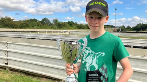 Jacob Kent in a green t-shirt and cap holds a trophy aloft next to a go-kart race track.