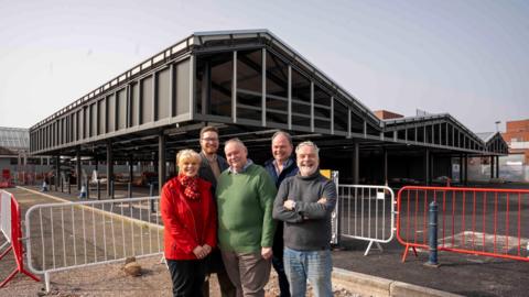 Four men and one woman stand in a group smiling at the camera. They stand in front of a construction site which is fenced off.