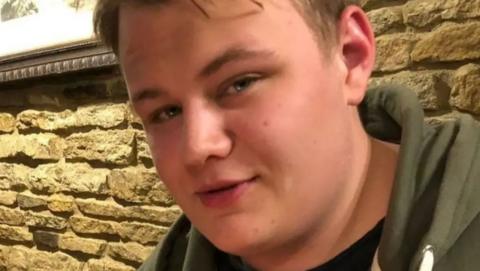 Close up of a young man's face. He is looking directly at the camera and is wearing a green hooded top.