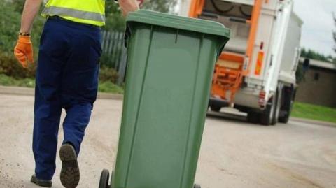 A binman carries a green recycling bin towards a recycling truck.