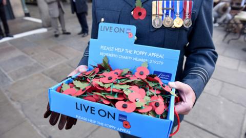 A veteran carrying a poppy appeal box with poppies inside it. The man is dressed in a blue uniform with a poppy and a number of medals. 