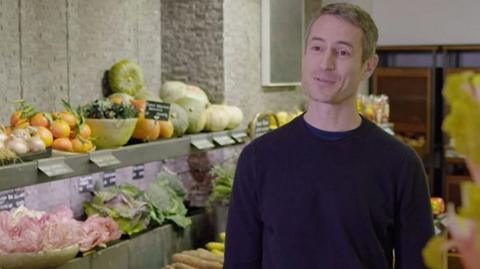 A man in a greengrocer shop with fruit and veg in the background.