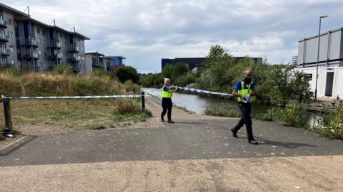 Two police officers roll out police tape to cordon off a waterway. Buildings and the water itself are visible behind them.