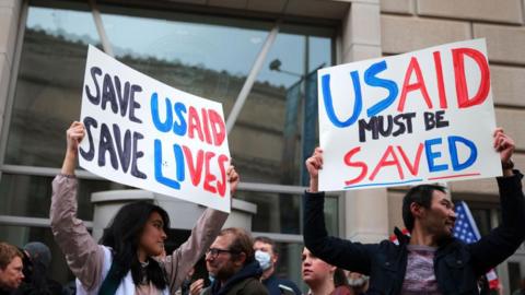 People holding protest signs in black, blue and red, reading: 'Save USAID, save lives' and 'USAID must be saved'