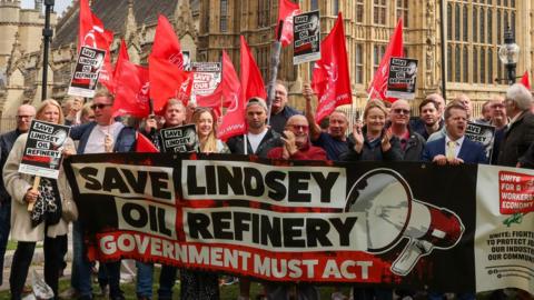 A group of more than 20 men and woman, some holding red flags and other holding black placards, stand in front of the Palace of Westminster and behind a large banner, coloured red, white and black, which reads: "Save Lindsey Oil Refinery" in large letters next to an image of a loud hailer. A second line of text on the banner reads: "Government must act".