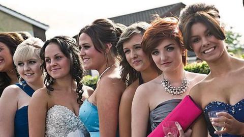 A group of female teenagers pose for a photo during a school prom. They all wear different prom dresses and smile at the camera. 