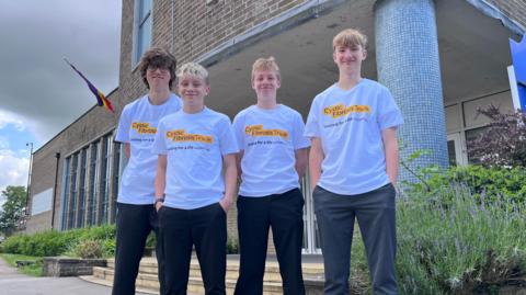 Four boys. Three have short blonde hair and one has slightly longer brown hair. They are smiling. Three have dark trousers and one has grey trousers. They are wearing white t-shirts with Cystic Fibrosis Trust written on them. They are stood in front of a brick building, a flag, tarmac and plants.