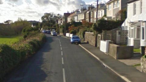 A country road , on the left is a field, and on the right are houses, some cars are parked on the road.