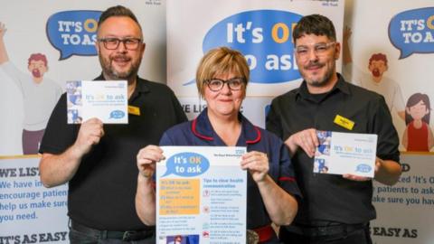 Two men with a woman in between them standing in front of tall, vertical banners that read "It's ok to ask". The woman is wearing a navy nurse's uniform with red piping and they are all wearing glasses. They are each holding a small poster that also reads "It's ok to ask" and they are smiling at the camera. 