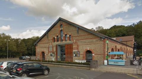 Entrance to Cheese and Grain which has decorative red bricks on building with a triangle top. There is a car park outside of the building.