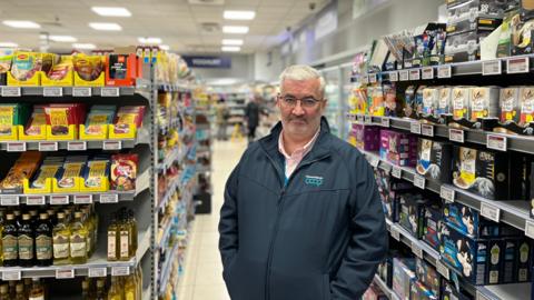 Mark Cox, a man with white hair, glasses and a grey beard. He is standing in a supermarket aisle, with pet food to the right of the image and cooking supplies - including olive oil and seasoning - to the left. He is wearing a 'coop' branded jacket with a zip through the middle, over a pink shirt.
