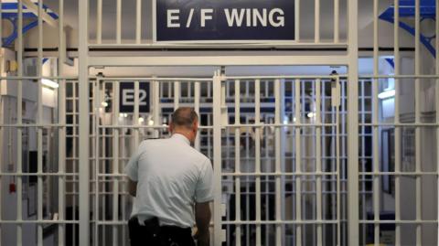 An image of a series of security gates in a prison - with floor-to-ceiling white metal bars. Above the first set it says "E/F Wing" and a prison officer - a bald man in a white shirt and black trousers - is opening the gate. We see him from behind.
