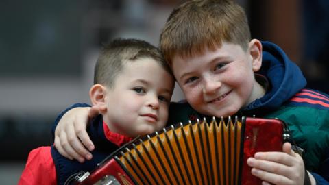 Two young boys with short brown hair sitting beside each other. They are holding an accordion.