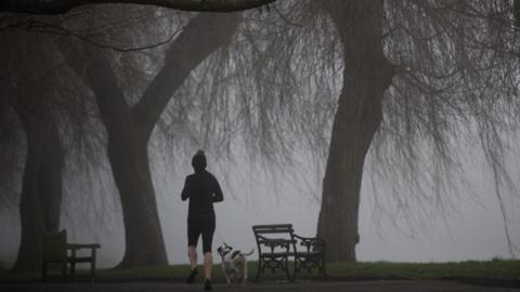 A woman in black running clothing runs with her dog in a misty and dark park.