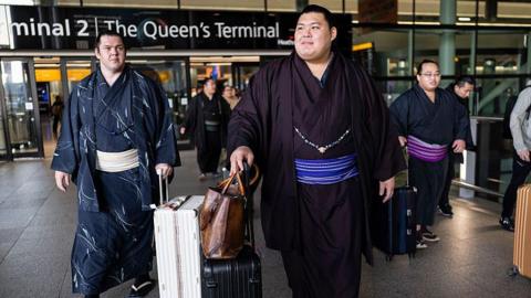 Three sumo wrestlers dressed in kimonos are walking and wheeling suitcases as they leave an airport.