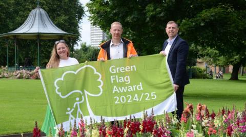 Three council officials holding a Green Flag Award in Mowbray Park. They are smiling and in the foreground there are purple and red flowers