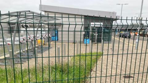 A fenced-off area outside a small modern station building in Winslow, Buckinghamshire, with bike racks on the left and ticket machines near the entrance.