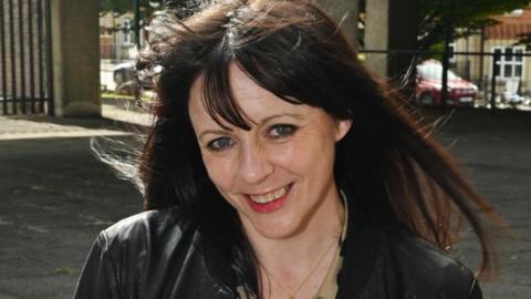 A close-up of Kathryn Olley. She has long, brown hair, is wearing a black leather jacket and is smiling.