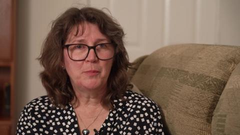 Lesley Ritchie is sitting a brown, patterned couch. She is wearing glasses and a white and white spotted top. 