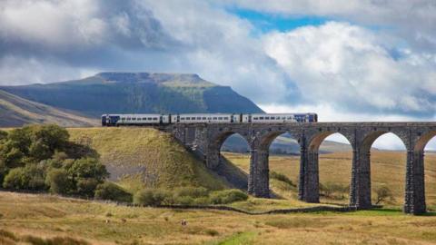 A Northern train crossing the Ribblehead Viaduct. The stone structure has several arches and is surrounded by open green space, with a fell behind it.