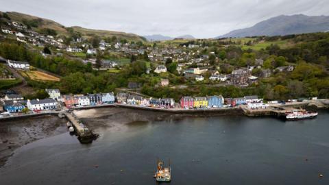 An aerial view of the Isle of Mull, including the port, with brightly coloured buildings at the sea front and sprawling hills in the distance