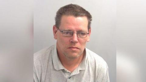 A police custody photo of a man in glasses who is wearing a grey polo shirt. He has short spiky hair.