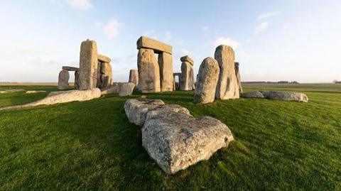 The Stonehenge circle of stones in the early morning light.  There is lots of green grass and there are shadows across some of the stones.