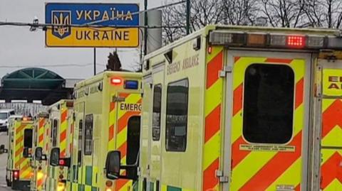 A line-up of five yellow London ambulances driving under a blue and yellow sign that says Ukraine, in both English and Ukrainian.  