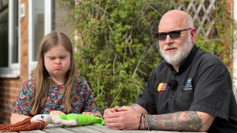 Image shows a girl wearing a flowery dress, playing with a mermaid doll. She has long light brown hair. On the right of the picture is a man with a black shirt, tattoos on his arm and dark glasses. He has a grey beard. In the background is a red brick house, with a green bush outside. 