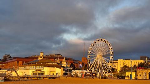 Dark moody skies create a backdrop for the early yellow sunshine illuminating the white seafront buildings and Ferris wheel at Bournemouth Pier with the beach in the foreground.