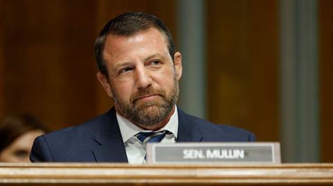 Mullin looks straight ahead and slightly smiles as he sits in a navy suit behind a placard of his name