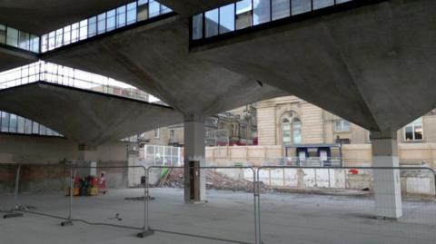 Grey concrete arches on top of pillars, with windows at the top as well. There are metal fences as bricks and work has started on the project.