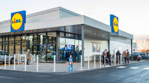 A Lidl store. It has a metal roof with signage outside and glass entrance with shoppers outside it.