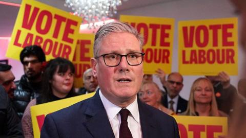 Sir Keir Starmer in a dark suit and dark tie and white shirt. He has grey hair and black rimmed glasses. There are yellow and red signs behind him which read Vote Labour and a crowd of people behind him. 