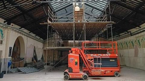 An orange machinery in front of scaffolding inside the empty Kendal Market Hall. There are ladders and building materials by the walls. 