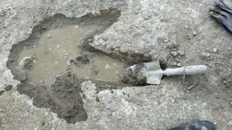 A dinosaur three toed footprint in grey mud, with the footprint filled with water. A trowel lies in the tip of the longest toe.
