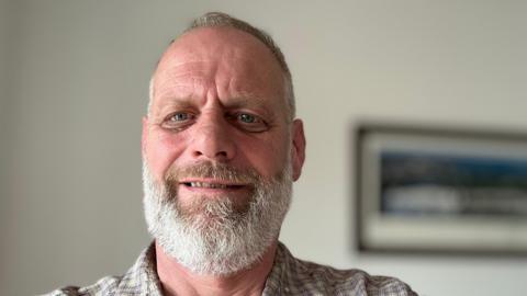 Simon Burke, a middle aged man with a white beard and hair. Wearing a blue checked shirt. 
