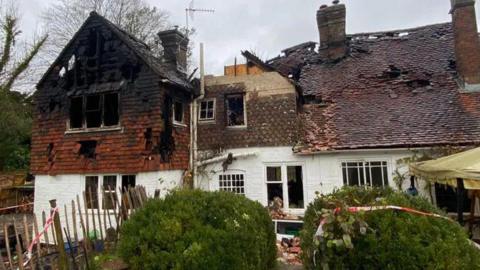 A house that has burned down, with holes in the roof and red and white safety tape around it and on the bushes at the front of the house. The sky in the background is grey and bushes can be seen on the left-hand-side.
