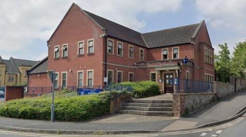 A brick building surrounded by blue railings with a hedge at the front partly obscuring a West Mercia Police sign