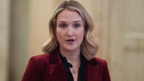 Helen McEntee has shoulder length blonde hair and is wearing a dark red blazer, with a black blouse underneath 