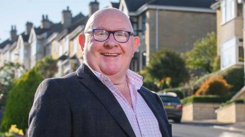 Rob Appleyard is standing in the sun in front of a row of houses. He is wearing a suit jacket over a pink checked shirt and is smiling at the camera.