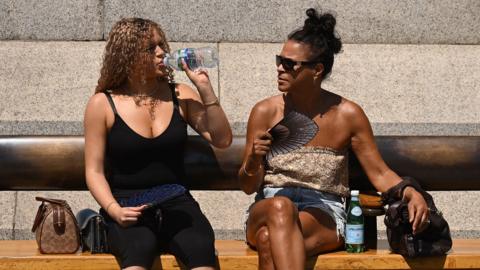 Two people sit down on a bench in Trafalgar Square to take some respite from the heat - one drinks a bottle of water and the other fans themself, in London on Friday