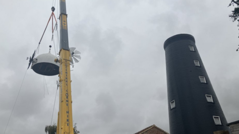 A yellow crane is lifting a white wooden mill cap back onto the top of a tall black mill. the sky is grey and cloudy