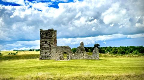 What appears to be ancient ruins of a building can be seen in a grassy field with trees in the far background. Clouds are gathering in the sky but areas of blue still remain