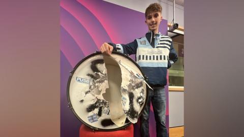 Cory stands next to his damaged drum. The skin is ripped from side to side, with the remnants showing the stickers fans have affixed to it over the years. Cory is wearing a Coventry City hooded top with 'Hummel' on the chest.