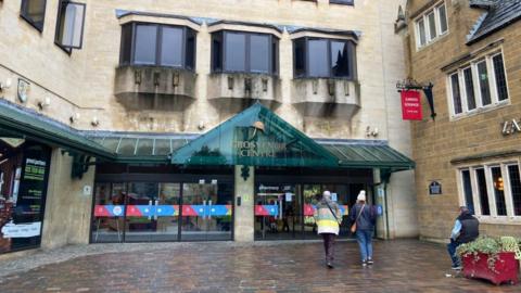 The front entrance to the Grosvenor shopping centre, featuring glass doors with green canopy. Triangular piece of canopy has the letters "Grosvenor Centre" in gold. Two people are heading towards the centre while a third is sitting outside a pub.