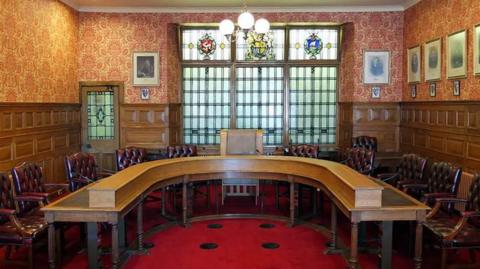 The Legislative Council chamber, which has brown wooden panelling on the walls and features a horseshoe-shaped table with maroon leather chairs around it on a red carpet. There is a stained-glass window at the back of  the  chamber with various crests on it. 