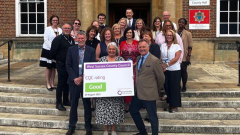21 people stood on the steps outside a brown brick council office building. They are smiling. One person at the front is holding a white board with the words West Sussex County Council CQC rating Good.